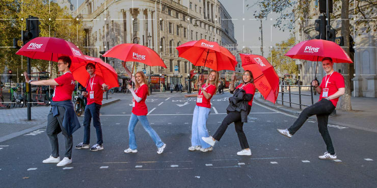 Six smiling Ping Identity team members holding branded red umbrellas while walking across a city street