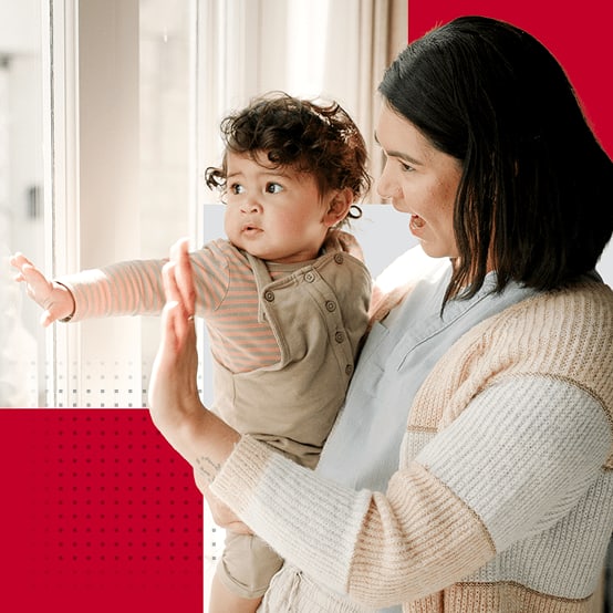 Female healthcare worker holding a toddler while waving through a window smiling
