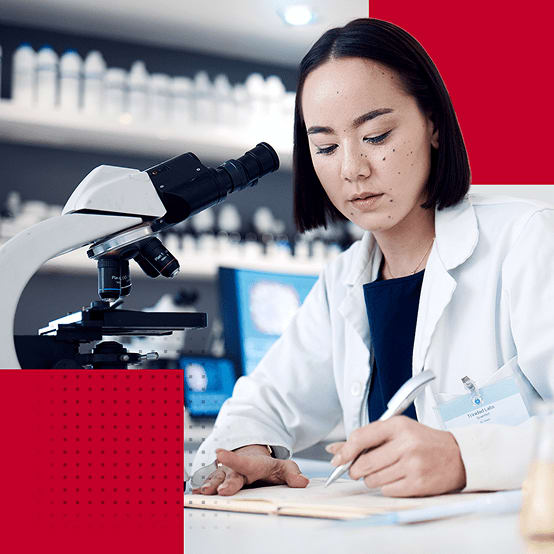 Woman in a lab coat writing notes beside a microscope